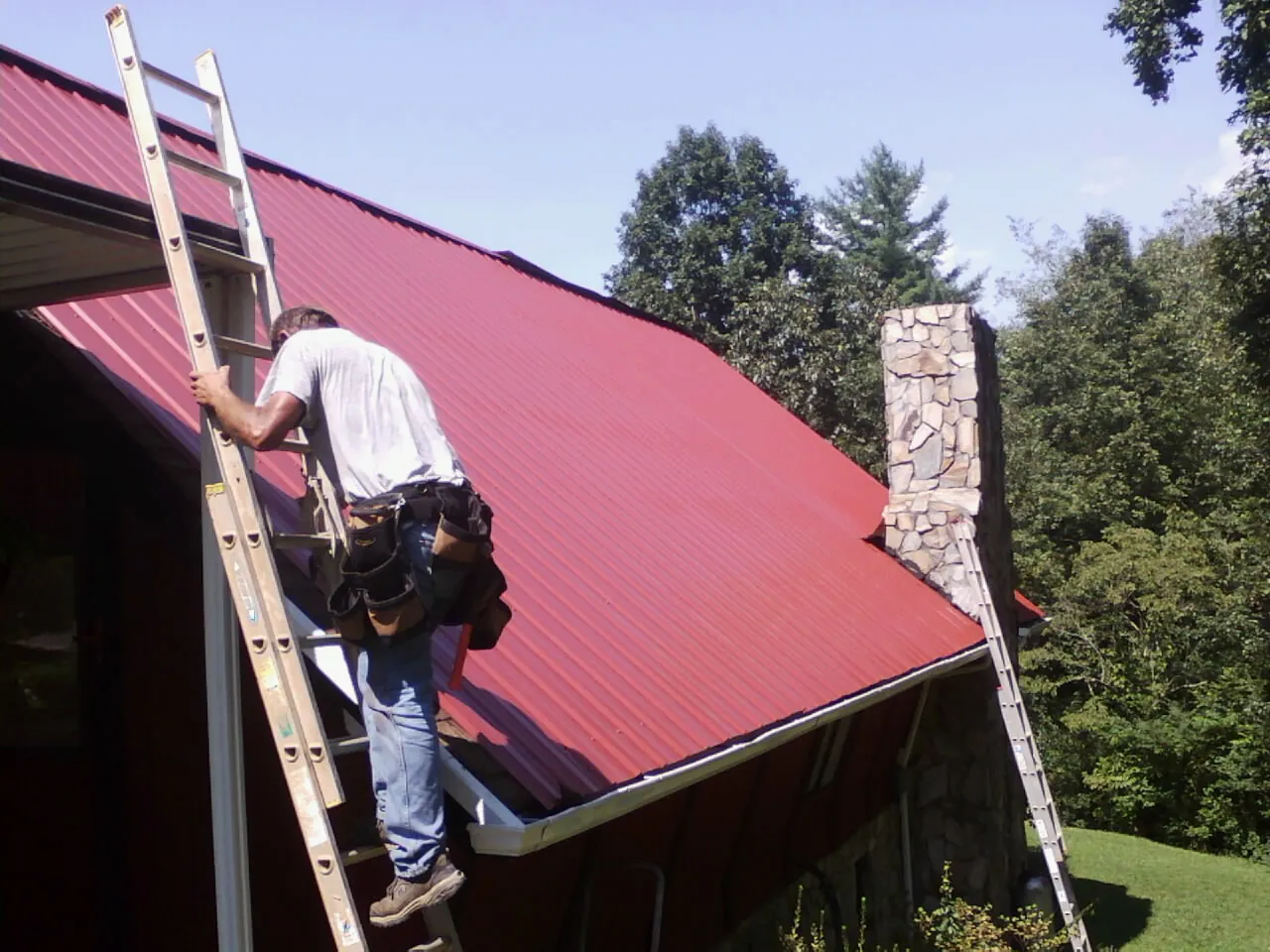 Roofer on a ladder beside a red metal roof and stone chimney — Soffit & Fascia Repair by Roofing Brothers in Cuttyhunk